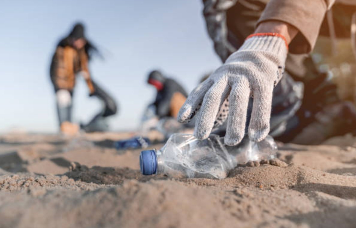 A gloved volunteer picking up a plastic bottle from the sand while others collect litter along the shoreline.