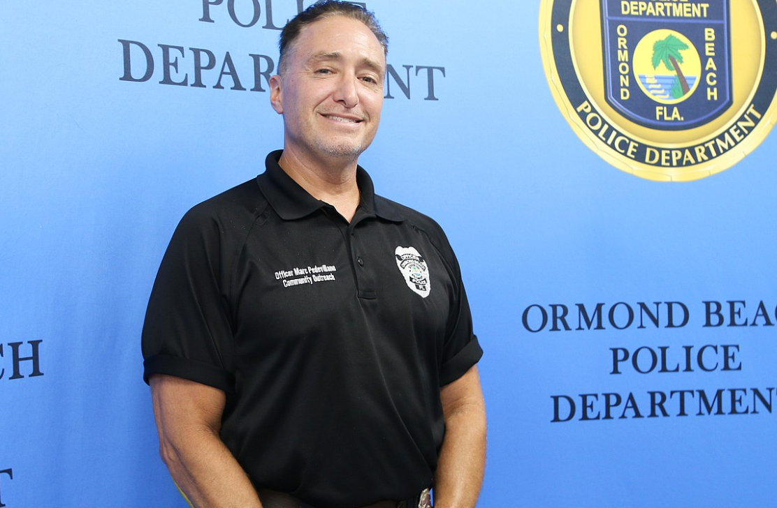 Ormond Beach Police Community Outreach Officer Marc Pedevillano standing in front of an Ormond Beach Police Department backdrop.